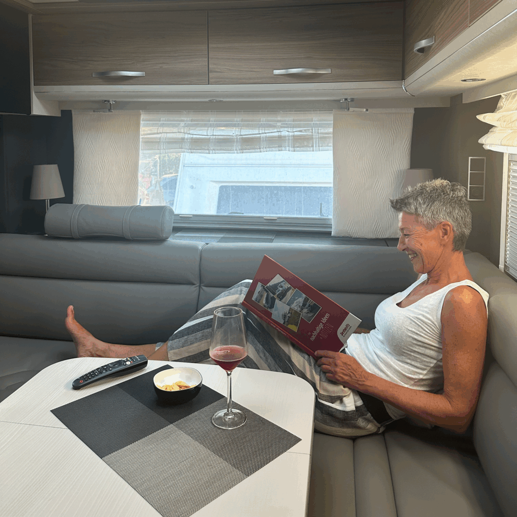 Frau liest entspannt ein Buch in der Sitzgruppe eines Dethleffs Alpa Wohnmobils, mit einem Glas Wein und Knabbereien auf dem Tisch.