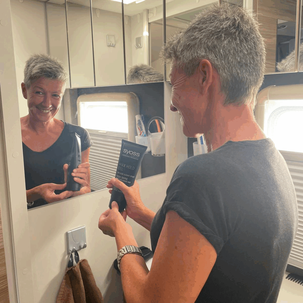 Frau hält Stylingprodukte vor dem Spiegel im Wohnmobil-Waschraum und lächelt in ihr Spiegelbild.